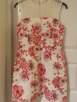 Vit och röd blommig kortklänning - Kort klänning med vit botten och stora röda blommor. Överdelen har spetsdetaljer i vitt och rund halsringning. Klänningen är ärmlös och har en figurnära passform. Perfekt för sommarens alla tillfällen.