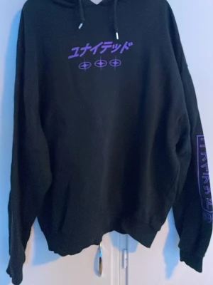 Svart hoodie med lila japansk text - Cool svart hoodie med huva och lila japansk text och grafik både fram och bak samt på ena ärmen. Dragsko i huvan och ribbade muddar. Perfekt för dig som gillar streetwear och unika tryck.
