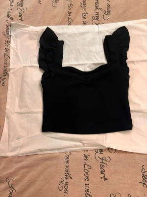 Svart crop top med volangärmar - Trendig svart crop top med breda axelband och volangdetaljer på ärmarna. Toppen har en fyrkantig urringning och är gjord i ett stretchigt, mjukt material som sitter snyggt på kroppen. Perfekt för att styla med höga jeans eller kjol.