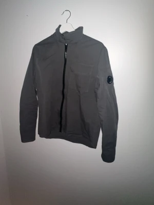 Grå vindjacka från C.P. Company - Säljer en grå vindjacka från C.P. Company med klassisk linsdetalj på ärmen. Jackan har hög krage, bröstficka och dragkedja framtill. Perfekt för dig som gillar stilrena och funktionella plagg.