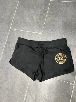 Svarta sportshorts med tryck - Snygga svarta lågmidjade sportshorts med guldfärgat tryck på ena benet där det står 'Grad Year Champions LTD 12 MMXII'. Shortsen har snörning i midjan och är tillverkade i ett lätt syntetmaterial som passar perfekt för träning eller bad.