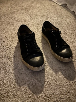 Svarta Lanvin sneakers med vita sulor - Snygga mörkblå sneakers från Lanvin med klassisk låg profil och vita gummisulor. Skorna har mörkblå skosnören och metallöljetter, samt en rundad tå i läder. Perfekta för dig som vill ha en stilren och exklusiv look. Jag har tyvärr inte något kvitto jag köpte dom i NK i Stockholm och köpte dom för 5400kr nya