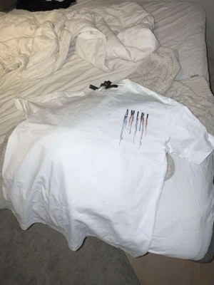 Vit AMIRI t-shirt med färgstänk - Säljer en vit t-shirt från AMIRI med tryck framtill och baktill där loggan har färgstänk i blått, orange och svart. T-shirten har korta ärmar och en klassisk passform. Perfekt för dig som gillar streetwear och vill sticka ut lite extra.