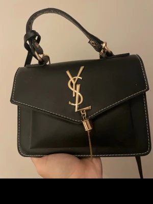 Svart Y handväska med gulddetaljer - Snygg svart handväska från Y i slätt läder med guldiga detaljer och logga framtill. Väskan har ett kort handtag och en bred axelrem med svartvitt mönster. Praktisk dragkedja på sidan och vit kontrastsöm runt kanterna.