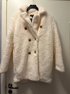 Vit teddyjacka från Eivy - Säljer en fluffig och vit teddyjacka med glansigt material på insidan. Jackan har dubbelknäppning med bruna knappar, hög krage och mjukt, lurvigt material.