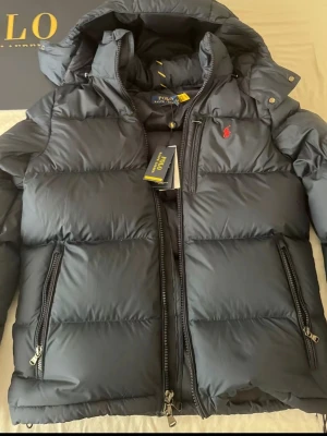 Svart pufferjacka från Polo Ralph Lauren - Snygg svart pufferjacka från Polo Ralph Lauren med röd broderad logga på bröstet. Jackan har huva, två dragkedjeförsedda fickor och tjock vaddering som håller dig varm. Klassisk design med hög krage och detaljer i metall. Perfekt för kalla vinterdagar.