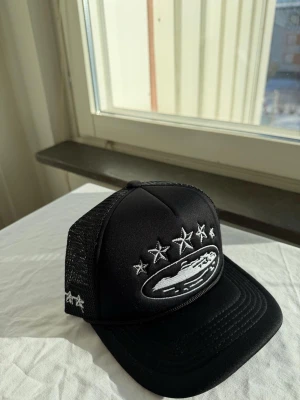 Svart truckerkeps med stjärnor - Snygg svart truckerkeps med mesh på sidorna och broderade vita stjärnor samt ett motiv av ett skepp framtill. Kepsen har böjd skärm och justerbar snapback baktill. Perfekt för en streetwear-look.