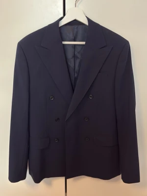 Mörkblå dubbelknäppt kavaj - Snygg mörkblå kavaj med dubbelknäppning och breda slag. Klassisk modell med två framfickor och innerfoder. Perfekt för en dressad look. Kavajen har fyra knappar framtill och är tillverkad i ett slätt tyg. Innerknappen sitter lite löst, därav priset. Men det är dock enkelt att fixa. Annars är kavajen i utmärkt skick.