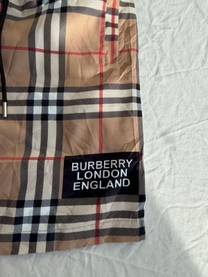 Rutig Burberry shorts med snörning - Säljer ett par klassiska Burberry shorts i beige med det ikoniska rutiga mönstret och svart snörning i midjan. Shortsen har en patch med 'BURBERRY LONDON ENGLAND' på benet och sidofickor. Perfekta för sommaren och riktigt snygga till streetwear.