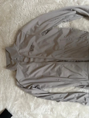 Beige vindjacka från EA7 Emporio Armani - Snygg beige vindjacka från EA7 Emporio Armani med diskret logga på bröstet. Jackan har dragkedja framtill, två sidofickor med dragkedja och en klassisk krage. Perfekt för dig som gillar stilrena och sportiga plagg.