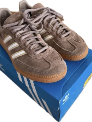 Bruna Adidas Spezial! - Säljer ett par bruna Adidas Spezial sneakers i mocka med klassiska vita och guldiga stripes på sidan. Skorna har gummisula och lågt skaft, samt matchande bruna skosnören. Perfekta för dig som gillar retrostil och streetwear.❣️❣️