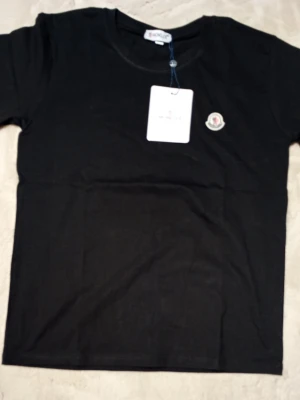 Svart t-shirt från Moncler - Säljer en svart t-shirt från Moncler med klassisk rund hals och korta ärmar. T-shirten har en snygg broderad Moncler-logga på bröstet och är tillverkad i mjuk bomull. Perfekt för dig som vill ha en stilren och exklusiv look. T-shirten passar som Large.