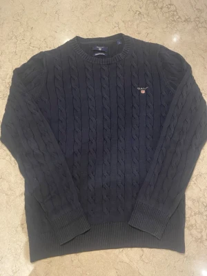 Mörkblå kabelstickad tröja Gant - Snygg mörkblå kabelstickad tröja från Gant i premium bomull. Tröjan har rund halsringning, ribbade muddar och Gant-logga broderad på bröstet. Perfekt för dig som gillar klassisk och stilren look.