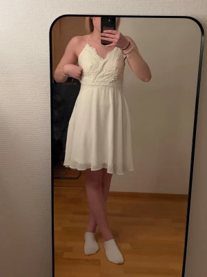 Vit spetsklänning med smala axelband - Från bubbelroom, oanvänd, Söt vit kortklänning med smala axelband och v-ringad spets upptill. Klänningen har ett blommigt spetsmönster på överdelen och ett lätt, flödande tyg i kjolen. 