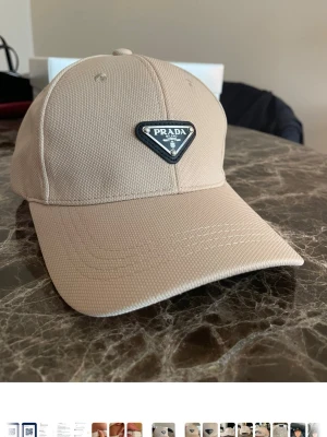 Beige keps helt ny  - Snygg beige keps från … med det ikoniska trekantiga metallmärket framtill. Kepsen har böjd skärm och är tillverkad i ett strukturerat tyg som ger en exklusiv känsla. Perfekt accessoar för dig som gillar stilrena och lyxiga detaljer.