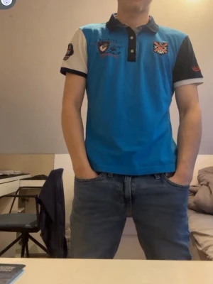 Blå polo t-shirt med patchar - Säljer en blå polo t-shirt från U.S. Polo Assn. med svarta och vita detaljer på ärmarna och kragen. T-shirten har coola patchar och broderier på bröstet och knappar vid halsen. Perfekt för dig som gillar en sportig och stilren look.