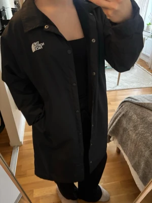 Svart coachjacka från The North Face - Snygg svart vindjacka från The North Face med tryckt logga på bröstet och stor logga på ryggen. Jackan har knäppning framtill, klassisk krage och en rak, längre passform. Fodrad med ”lurv” inuti