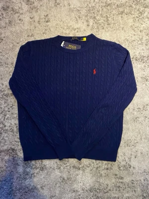 Mörkblå Ralph Lauren tröja  - Säljer denna klassiska kabelstickade Ralph Lauren tröjan i mörkblå med röd logga. Storleken är L fast passar mer som en mindre M enligt mig, tröjan är dessutom i mycket fint skick. Hör gärna av dig vid frågor om t.ex. mått, bilder etc!