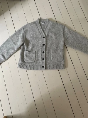 Grå stickad kofta från Weekday - Mysig grå stickad kofta från Weekday med v-ringning, svarta knappar och två stora fickor framtill. Koftan har ribbade muddar vid ärmslut och nederkant samt en avslappnad passform. Perfekt för lager på lager och enkel att styla. Har använt 2 gånger💕skriv för fler bilder!💕