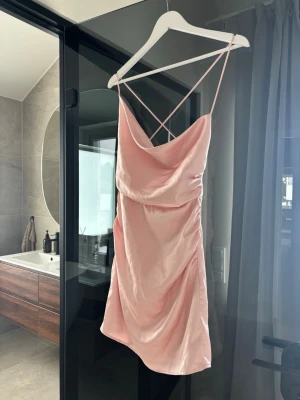 Ljusrosa kort satinklänning - Supersöt ljusrosa kort klänning i satin med tunna axelband som korsas i ryggen. Klänningen har en draperad urringning och en figurnära passform som ger en elegant look. 