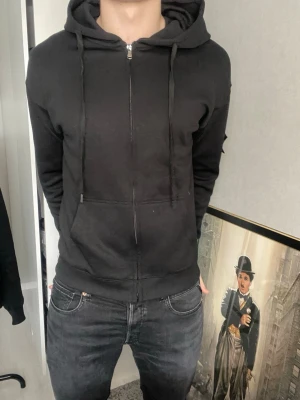Svart hoodie från Stone Island - Säljer en svart hoodie från Stone Island med dragkedja och huva. Klassisk logopatch på vänster ärm och stora fickor framtill. Perfekt för dig som gillar streetwear och vill ha en stilren look. Materialet är mjukt och bekvämt, med ribbade muddar och snören i huvan. Tröjan är i stolek M! 