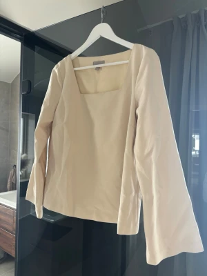 Beige blus med fyrkantig urringning - Stilren beige blus med lång ärm och trendig fyrkantig urringning. Blusen har en rak passform och är tillverkad i ett mjukt, lätt glansigt tyg som ger en elegant känsla.