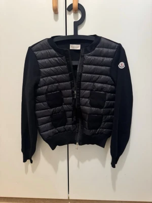 Moncler cardigan - Snygg svart cardiganjacka från Moncler med quiltad framsida och två fickor. Jackan har rund halsringning, ribbade muddar och dragkedja framtill. Ärmarna är stickade och vänster ärm har Moncler-logga. Perfekt för lager på lager och streetstyle. Kvitto finns