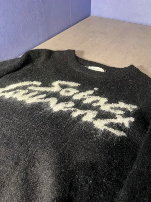 Svart stickad tröja från Saint Laurent - Cool svart stickad tröja från Saint Laurent med vit text på bröstet. Tröjan har en rund halsringning och är gjord i ett fluffigt material som ger en trendig look. Perfekt för dig som gillar statement-plagg och vill sticka ut.