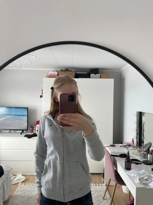 Grå zip-hoodie från Puma - Säljer en grå hoodie från Puma med dragkedja och huva. Tröjan har ett svart Puma-logo på bröstet, ribbade muddar och fickor framtill. Perfekt för chill dagar och sportiga outfits. Materialet är mjukt och bekvämt, troligen bomullsblandning.