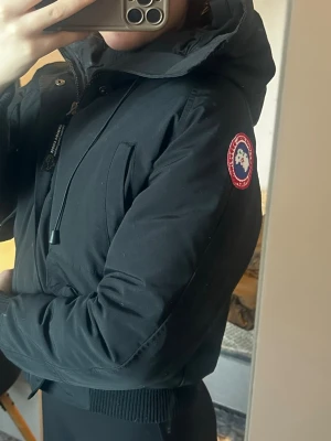 Svart Chilliwack bomberjacka Canada Goose - Säljer en svart Chilliwack bomberjacka från Canada Goose med ikonisk patch på ärmen, dragkedja och knappar framtill, flera fickor och ribbade muddar. Jackan har en stor huva med dragsko och pälsdetalj. Perfekt för kalla dagar och riktigt snygg streetstil.