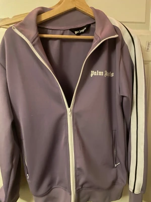 Lila ziptröja från Palm Angels - Säljer en lila ziptröja från Palm Angels med vita detaljer och svarta ränder på ärmarna. Tröjan har hög krage, dragkedja framtill och fickor med dragkedja. Materialet är en mjuk blandning av bomull och polyester. Perfekt för en sportig och trendig look.