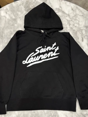 Svart Saint Laurent hoodie med tryck - Snygg svart hoodie från Saint Laurent med stort vitt logotyp-tryck på bröstet. Klassisk känguruficka framtill, dragsko i huvan och ribbade muddar vid ärmar och nederkant. Perfekt för en chill och trendig look.