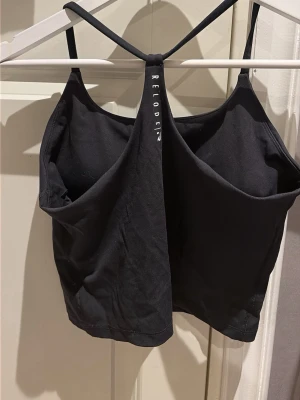 Svart croppad linnetopp Relode - Svart croppad linnetopp från Relode med tunna axelband och öppen rygg. Toppen har en stilren design med logga på ryggen och är tillverkad i mjuk bomullsmix som känns skön mot huden. Perfekt för sommarens alla outfits.