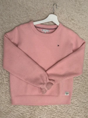 Rosa stickad tröja Tommy Hilfiger - Mysig rosa stickad tröja från Tommy Hilfiger med rund halsringning och diskret logga på bröstet. Tröjan har lång ärm och ribbade muddar vid ärmslut och nederkant. Perfekt för lager-på-lager och chill vardagsstil.