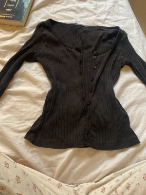 Svart ribbad långärmad kofta - Brandy Melville Svart ribbad kofta med lång ärm och knappar framtill. Koftan har en djup rundad halsringning och är tillverkad i ett stretchigt material som sitter snyggt på kroppen. Perfekt för att skapa en chill och stilren look.