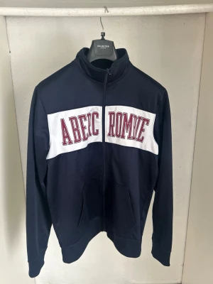 Mörkblå ziptröja Abercrombie - Mörkblå ziptröja från Abercrombie med vit panel och röd text på bröstet. Tröjan har hög krage, hel dragkedja och två fickor framtill. Tillverkad i mjukt material, perfekt för en avslappnad och sportig stil.