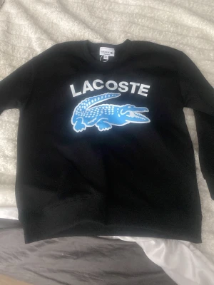 Svart sweatshirt från Lacoste - Säljer en svart sweatshirt från Lacoste med stort blått och vitt krokodilmotiv och texten 'LACOSTE' på bröstet. Tröjan har rund halsringning och långa ärmar. Perfekt för dig som gillar streetwear och vill ha en snygg statement-piece. M men kan passa S lika gärna. Helt ny oanvänd. 