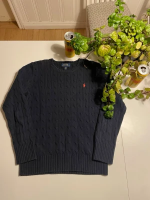 Mörkblå kabelstickad tröja Polo Ralph Lauren - Mörkblå kabelstickad tröja från Polo Ralph Lauren med klassisk rund halsringning och röd broderad logga på bröstet. Tröjan har ribbade muddar vid ärmslut och nederkant. Perfekt för en stilren och avslappnad look.