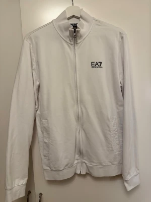 Vit zip-hoodie från EA7 Emporio Armani - Snygg vit hoodie med dragkedja från EA7 Emporio Armani. Stilren design med logga på bröstet och diskreta ränder i nacken. Mjuk bomullskänsla, två fickor fram och ribbade muddar. Perfekt för en clean och sportig look. Passar M även 
