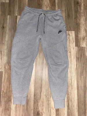 Gråa mjukisbyxor från Nike - Säljer ett par gråa mjukisbyxor från Nike med svart logga på vänster ben. Byxorna har dragsko i midjan, muddar vid bensluten och en praktisk ficka med dragkedja bak. Tillverkade i mjukt sweatshirtmaterial för maximal komfort.