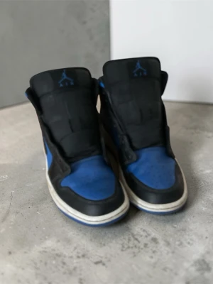 Nike Air Jordan 1 Mid Svart/Blå - Snygga Nike Air Jordan 1 Mid sneakers i svart och blått läder med klassisk swoosh på sidan och blå detaljer på tungan. Utan snören. Skorna har en vit sula med blå yttersula och ikonisk high-top siluett. Perfekta för dig som gillar streetwear och basketstil. Hör av er för frågor! Och kan sänkas en bit. Med vänlig hälsning, Valter ☺️