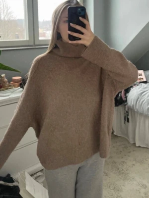 Oversize brun polotröja stickad - Mysig och oversize stickad polotröja i en varm brun nyans. Tröjan har långa ärmar och hög krage som ger en avslappnad look. Perfekt att slänga på sig när det är kyligt ute. Materialet ser ut att vara mjukt och fluffigt.