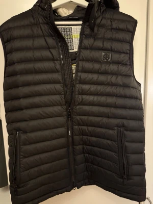 Svart dunväst med huva från moose knuckles  - Snygg svart dunväst från moose knuckles med horisontella sömmar. Västen har två dragkedjeförsedda fickor fram och en diskret logotyp på bröstet. Perfekt för lager-på-lager och håller dig varm under kyliga dagar. Knappt använt den bra skick.