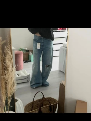 Blåa vida jeans med slitningar - Säljer ett par ljusblå jeans med bred passform och slitningar på ena benet. Slutsålda!