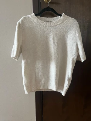 Vit stickad t-shirt med kort ärm - Säljer en enkel och stilren vit stickad t-shirt med korta ärmar och rund halsringning. Tröjan har ribbade muddar vid hals och nederkant. Säljer för att jag har växt ur den. Från H&M. Storlek S.