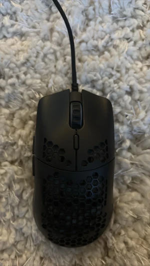 Glorius model 0 mini - Säljer en svart gamingmus med honeycomb-design och sladd. Musen har ett lättviktigt plastskal med hexagonmönster för bättre ventilation och grepp. Perfekt för gaming och snabba rörelser. Ergonomisk form och scrollhjul på toppen.