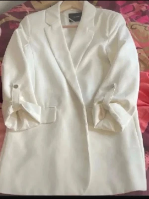 Vit kavaj med uppvikta ärmar - Stilren vit kavaj från Vila med klassisk krage och uppvikta ärmar. Kavajen har två fickor framtill och en rak passform som ger en snygg siluett. Perfekt för att lyfta vilken outfit som helst.