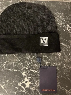 Svart mössa från Louis Vuitton - Snygg svart mössa från Louis Vuitton med diskret rutmönster och vikt kant. Märkt med LV-logga framtill på en vit patch. Tillverkad i mjukt stickat material som känns lyxigt och bekvämt. Perfekt accessoar för att höja din apart Edel-stil.