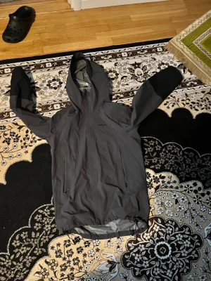 Mörkgrå regnjacka från Peak Performance - Snygg mörkgrå regnjacka med huva och två stora dragkedjeförsedda fickor framtill. Jackan har en minimalistisk design och är tillverkad i ett vattenavvisande material. Perfekt för regniga dagar och enkel att packa ner i väskan.