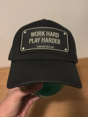 Svart truckerkeps John Hatter & Co - Snygg svart truckerkeps från John Hatter & Co med mesh på sidorna och bak. Framtill sitter en stor patch med texten 'WORK HARD PLAY HARDER'. Klassisk böjd skärm och justerbar passform. Perfekt statement piece för dig som gillar streetwear.